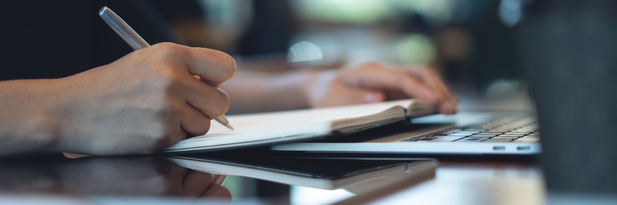 Mãos anotando em caderno ao lado de tablet e teclado de notebook