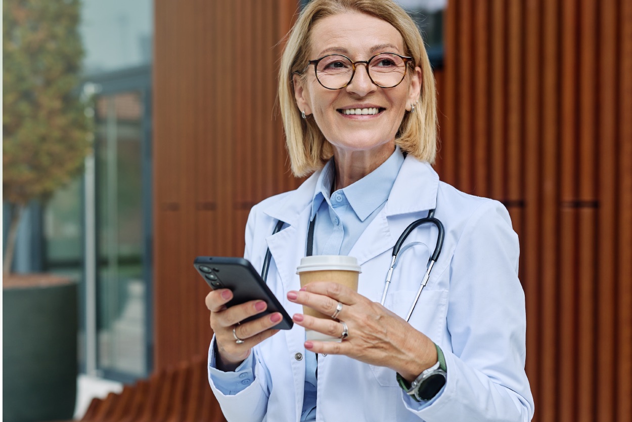 Médica sorridente de jaleco e estetoscópio segurando celular e copo de café