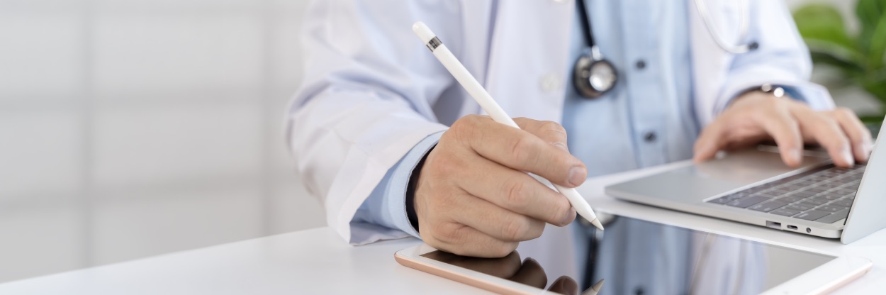 Profissional de jaleco branco com estetoscópio escrevendo em tablet ao lado de um notebook